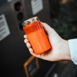 Person hält Grabkerze in rotem recyceltem Mehrwegbecher in der Hand. Vor einem Grablicht Service Point der Firma Vater & Sohn Grablicht GmbH