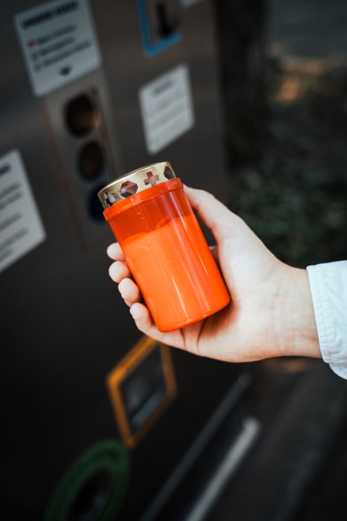 Person hält Grabkerze in rotem recyceltem Mehrwegbecher in der Hand. Vor einem Grablicht Service Point der Firma Vater & Sohn Grablicht GmbH